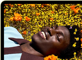 MacBook Neo, color amarillo cítrico, la pantalla muestra a una persona sonriente acostada en un campo de flores bajo el sol